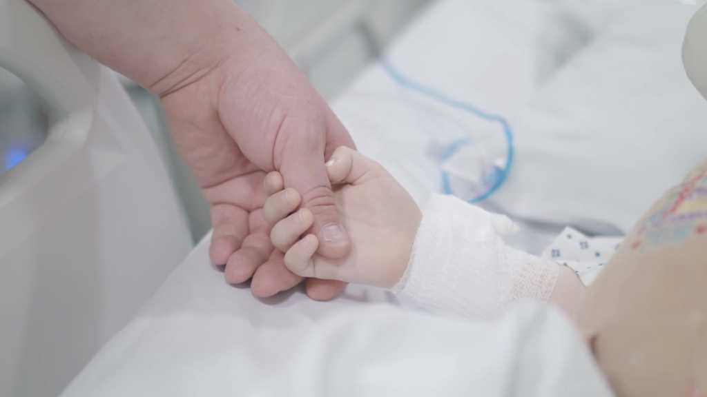 Adult hand holding a baby's hand in a hospital bed